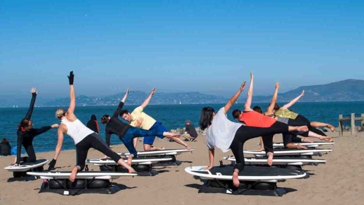 A beach yoga group class on static surfboards on the sand. women are doing a triangle pose with a blue sea and sky in the background