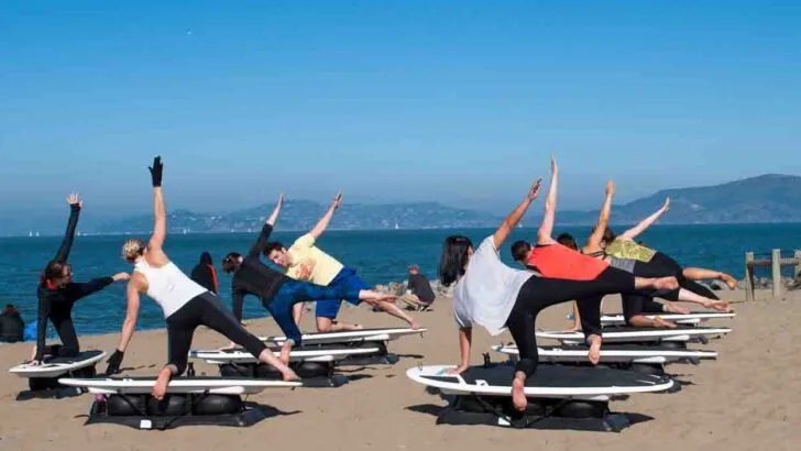 A beach yoga group class on static surfboards on the sand. women are doing a triangle pose with a blue sea and sky in the background
