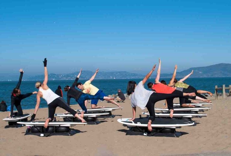 A beach yoga group class on static surfboards on the sand. women are doing a triangle pose with a blue sea and sky in the background
