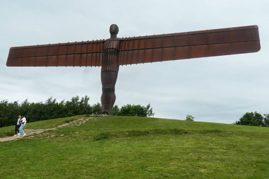 Angel of the North. Sculpture by Antony Gormley. The statue stands on a grassy hill with two people walking down a path in front of it. There are trees and shrubs to the left of it.