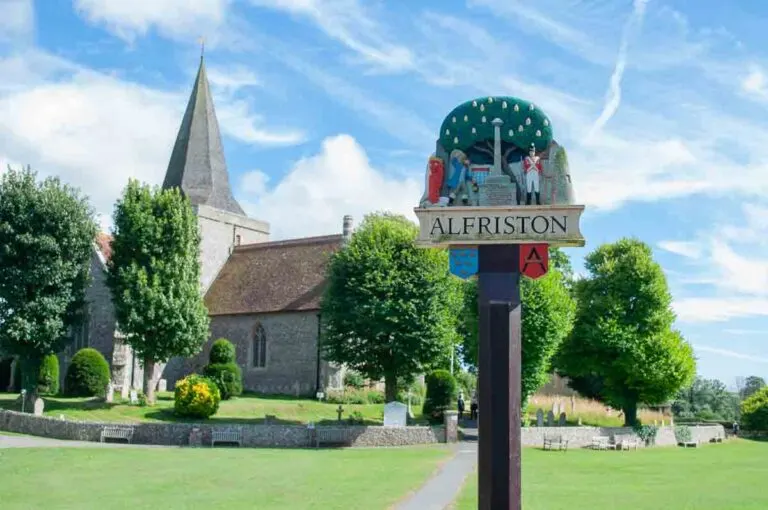 The Tye, Alfriston village in East Sussex. A village sign for Alfriston, England, stands in a green churchyard with a historic stone church.