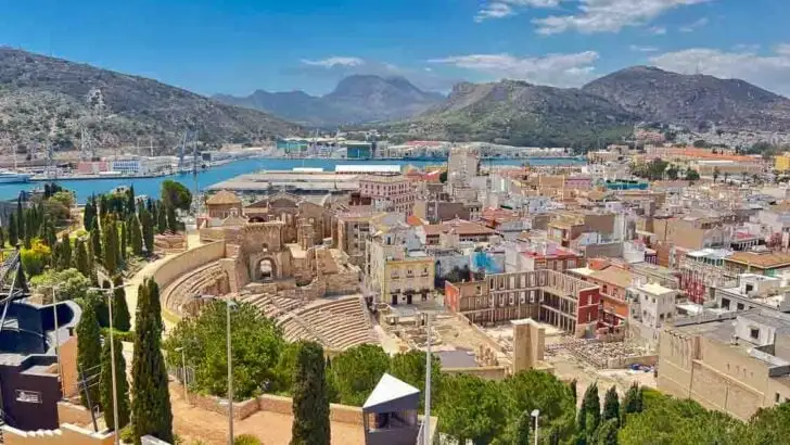 One of the best things to do in Cartagena Spain is to take in this view of the ancient Roman Theatre in Cartagena, Spain, showcasing its semi-circular seating carved into the hillside, with remnants of the stage and surrounding structures under a clear blue sky.