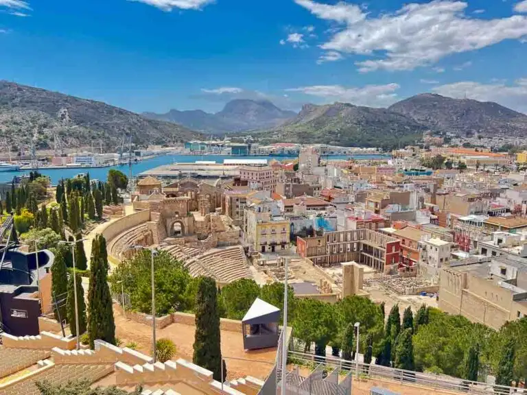 One of the best things to do in Cartagena Spain is to take in this view of the ancient Roman Theatre in Cartagena, Spain, showcasing its semi-circular seating carved into the hillside, with remnants of the stage and surrounding structures under a clear blue sky.