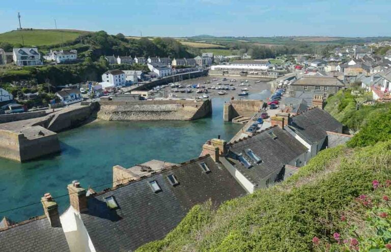 Looking down on Porthleven Harbour over a row of rooftops. Green hills are in the background and the blue sea waters sits within the thick harbour walls