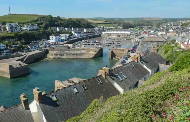 Looking down on Porthleven Harbour over a row of rooftops. Green hills are in the background and the blue sea waters sits within the thick harbour walls