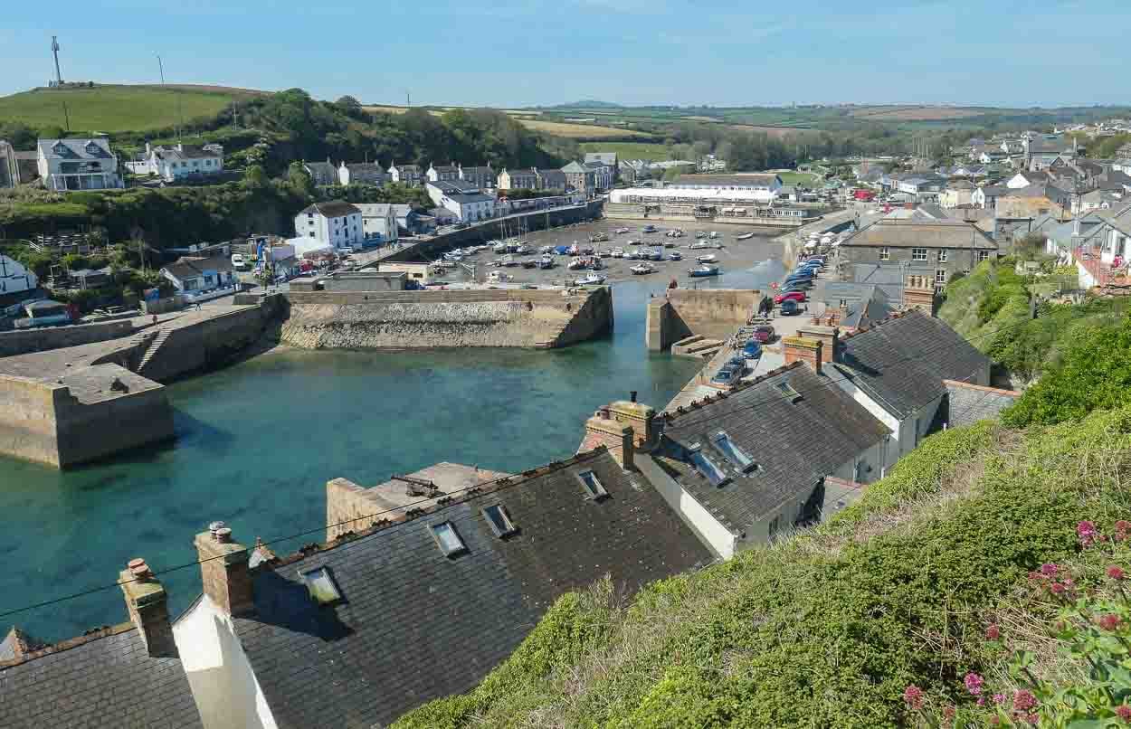 Looking down on Porthleven Harbour over a row of rooftops. Green hills are in the background and the blue sea waters sits within the thick harbour walls