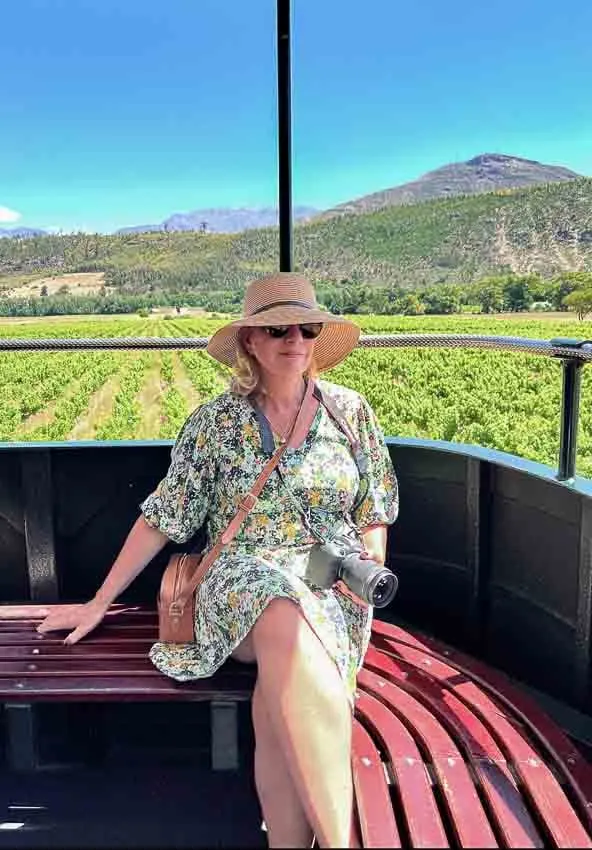 Woman with hat and sunglasses sitting on the top deck of the Franschhoek wine tram with mountains and vineyard in the background