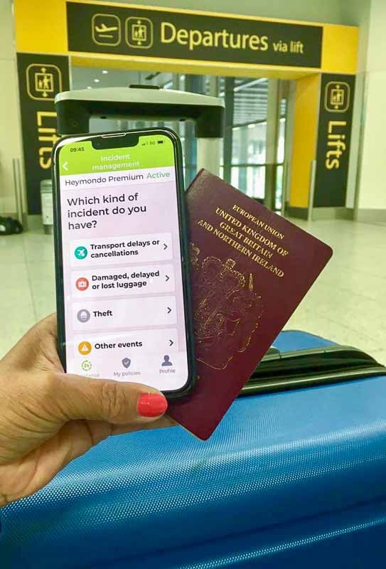 womans hand hold mobile phone with travel insurance app and passport in front of an airport departures sign. A blue suitcase is in the background