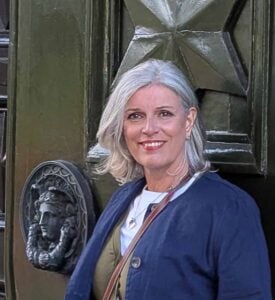 Suzanne Jones aka The Travelbunny UK travel blogger. Woman with shoulder length silver hair is smiling at camera and standing in front of an olive green door with a large black lion door knocker. She is wearing a white t-shirt with an olive green waistcoat and a navy jacket