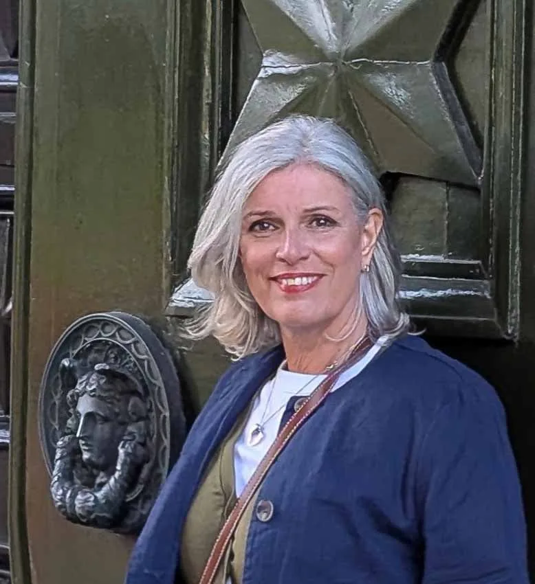 Suzanne Jones, travel blogger and content creator. Middle-aged woman with shoulder lenghth grey hair standing in front of an old green door with a large black knockers. She is smiling at camera and wearing a dark blue jacket, white t-shirt and olive green waistcoat.
