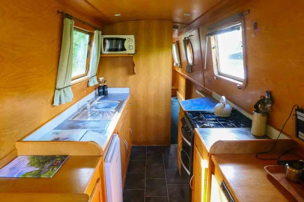 Narrow boat galley with worktop, sink and microwave on the left with a window above and pretty green curtains. On the right hand side is a small porthole window, gas hob and cooker with a kettle on top