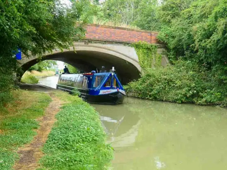 A. blue narrow boat is coming towards the viewer under an arched brick bridge. Trees are on either side of the canal with the grassy towpath in the foreground