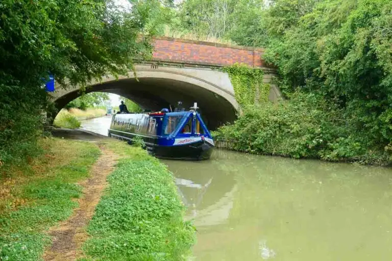 A. blue narrow boat is coming towards the viewer under an arched brick bridge. Trees are on either side of the canal with the grassy towpath in the foreground