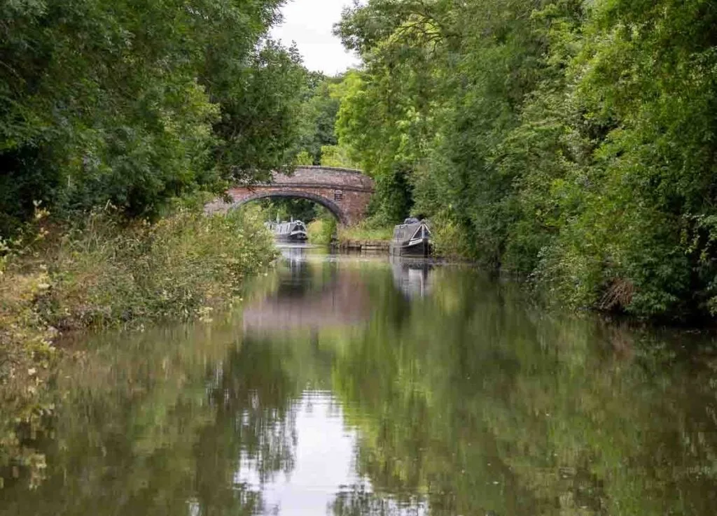 Narrow boat holidays. An English canal lined with trees which are reflected in the water. Two narrow boats are moored with a red brick arched bridge separating them