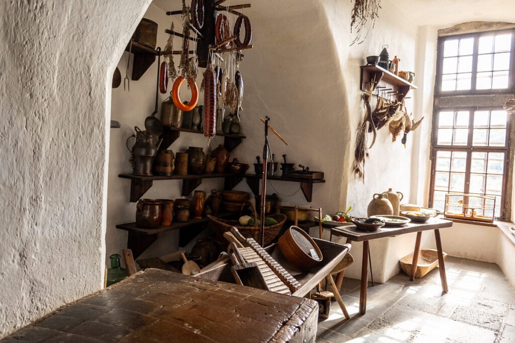 Marksburg Castle kitchen. A range of medieval cooking implements sit in an alcove. A platter of vegetables sit on a table and light floods in through the window
