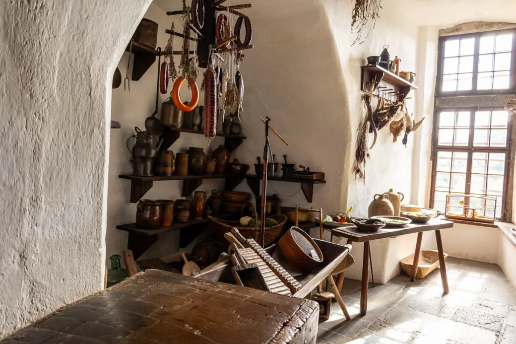 Marksburg Castle kitchen. A range of medieval cooking implements sit in an alcove. A platter of vegetables sit on a table and light floods in through the window
