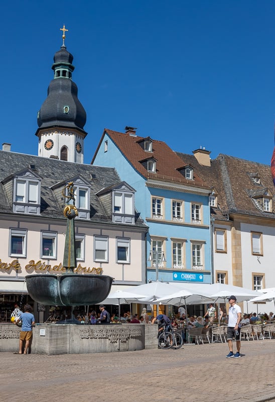 Speyer town centre, Germany. Old pastel coloured building stand behind a fountain with the spire of a church behind them and a blue sky
