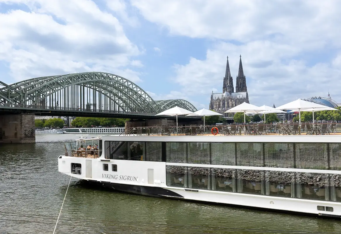 Viking ship Sigrun sits on the Rhine. There are parasols on the top deck with tables and chairs. At the bow of the ship people are dining. In the background is Cologne's Hohenzollern Bridge to the left and the city's twin towers of the cathedral peeping out behind the boat.