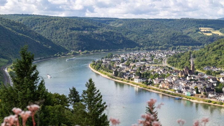 View of the Rhine River from Marksburg Castle looking down on a bend in the river with a tree-lined bank to the right and a village lining the river to the right. White fluffy clouds are in the background