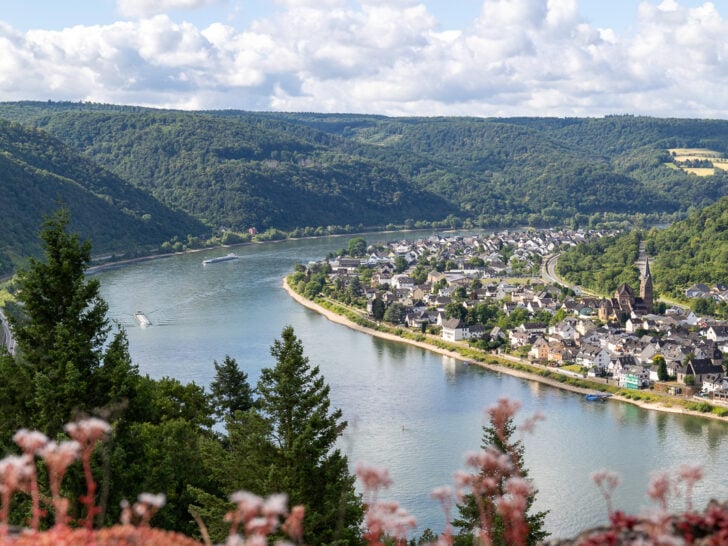 View of the Rhine River from Marksburg Castle looking down on a bend in the river with a tree-lined bank to the right and a village lining the river to the right. White fluffy clouds are in the background