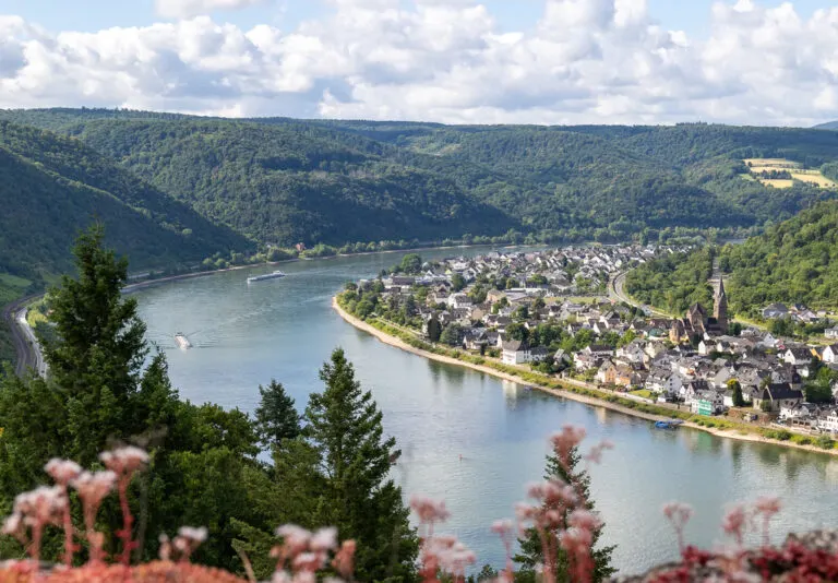 View of the Rhine River from Marksburg Castle looking down on a bend in the river with a tree-lined bank to the right and a village lining the river to the right. White fluffy clouds are in the background