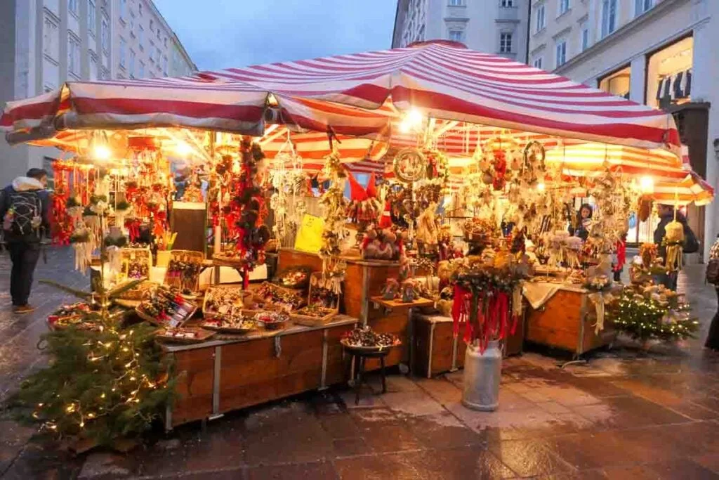 Christmas stalls in the Alter Markt, Salzburg. Under a red and white striped parasols are Christmas foods, gifts and small lit Christmas trees. Lights shine down from the parasols giving everything a warm yellow glow. It's raining