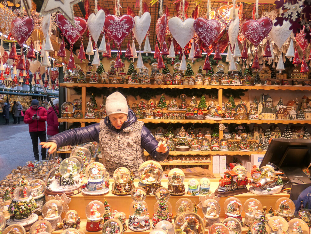 A stall at Salzburg Christmas loaded with Christmas baubles, decorations and snow globes. Tin hearts and Christmas trees are hanging from the ceiling. A woman in a white beanie is serving.
