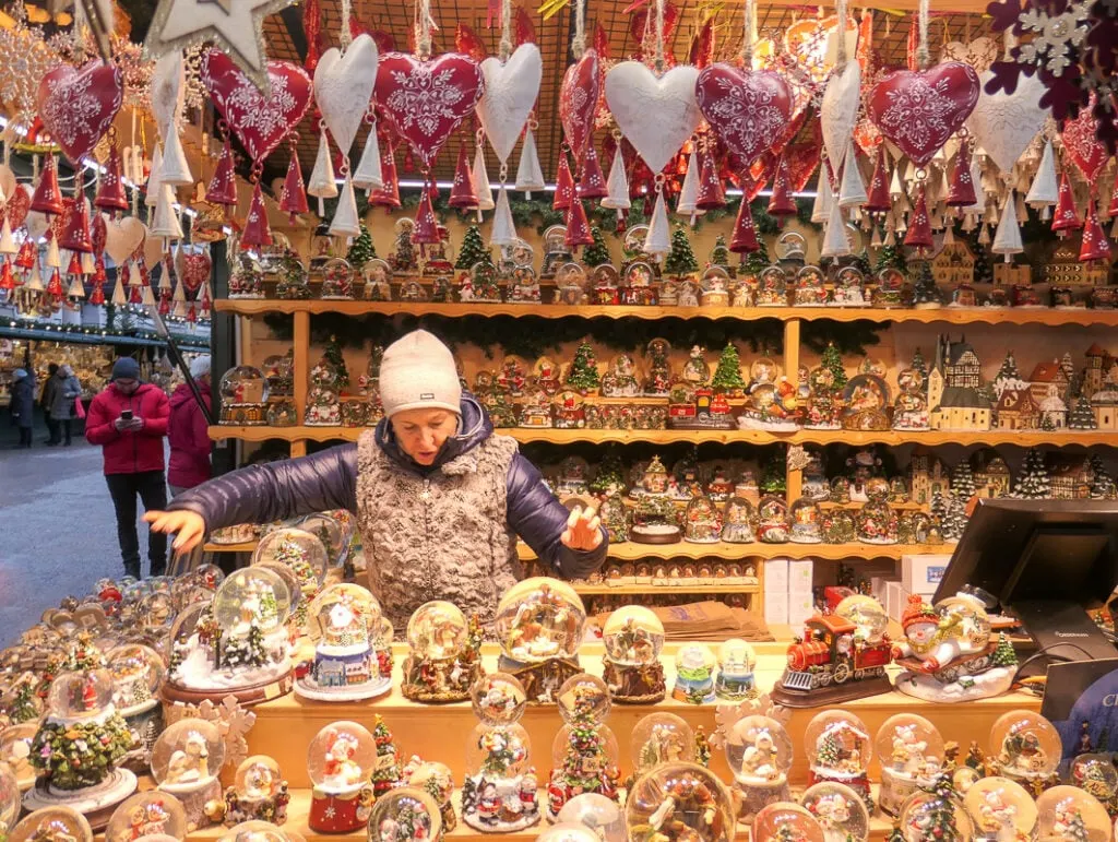 A stall at Salzburg Christmas loaded with Christmas baubles, decorations and snow globes. Tin hearts and Christmas trees are hanging from the ceiling. A woman in a white beanie is serving.