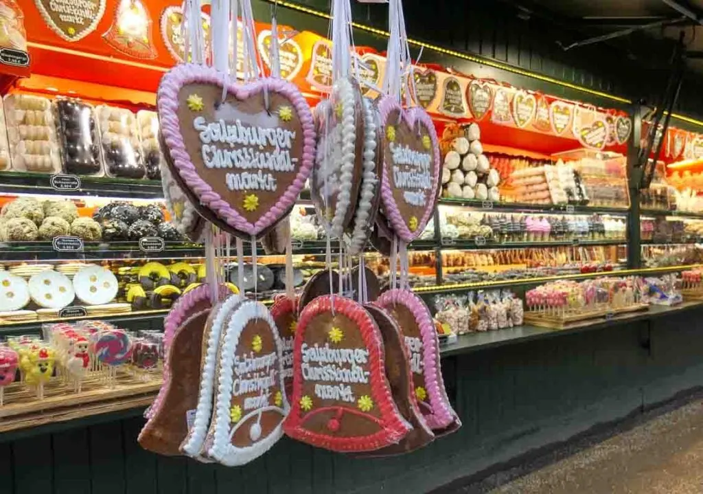 A Christmas market stall in Salzburg with rows of sweets, cakes, cookies and lollipops for sale. In the foreground, hanging from the front of the stall, are christmas gingerbread cookies shaped like bells and hearts with the words Salzburge Christmarkt piped in icing on them