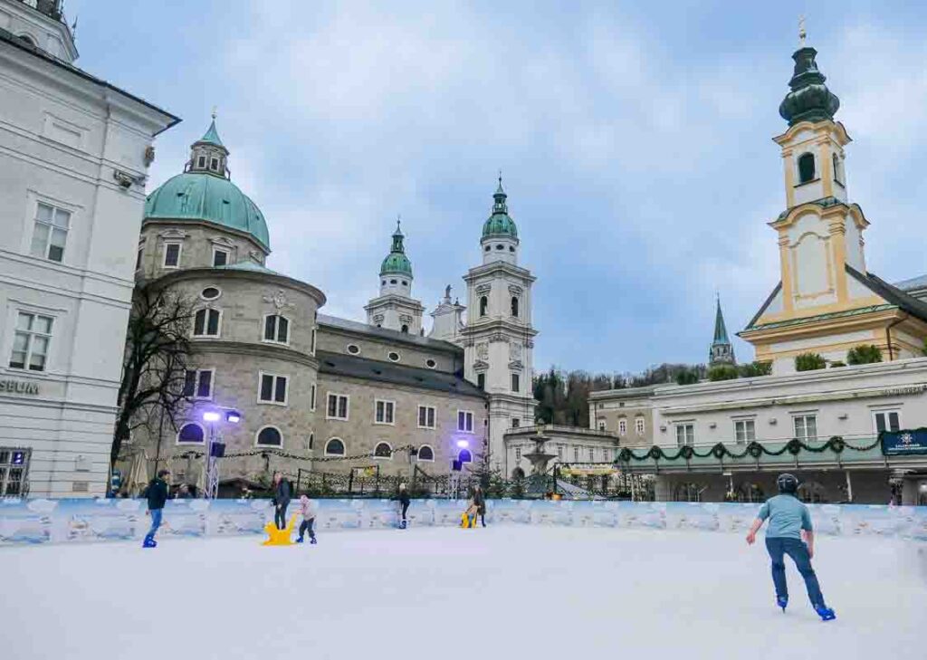 Eiszauber ice skating rink in Salzburg. The cathedral can bee seen in the background. It's a grey day and a man is skating wearing a helmet and grey t-shirt. Two children are on the ice with yellow supports and three adults clutch the side walls