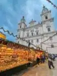 Christmas stalls in Salzburg, Austria. Brightly lit stalls are loaded with sweets, cookies and confectionery. The cathedral is in the background and green garlands with fairy light a strung above the stalls. People are browsing at the goods
