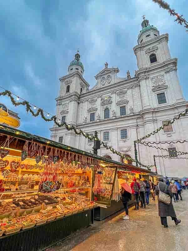Christmas stalls in Salzburg, Austria. Brightly lit stalls are loaded with sweets, cookies and confectionery. The cathedral is in the background and green garlands with fairy light a strung above the stalls. People are browsing at the goods