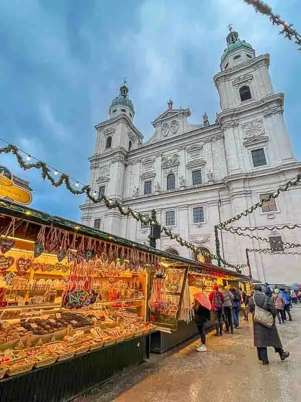 Christmas stalls in Salzburg, Austria. Brightly lit stalls are loaded with sweets, cookies and confectionery. The cathedral is in the background and green garlands with fairy light a strung above the stalls. People are browsing at the goods