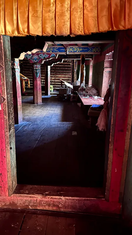 Inside the wooden Dege Parkhang Sutra-Printing House. Looking through a doorway into a room with carved, painted beans with desk sitting under light coming in throught he windows
