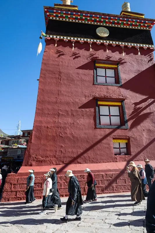 Dege Parkhang Sutri Print House with shadows cast across the dark red traditional building and people wearing hats walking in front of it