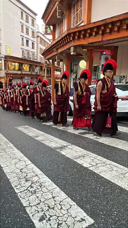 A row of dozens Tibetan monks in maroon robes and red headresses walking up a road in Dege, Sichuan
