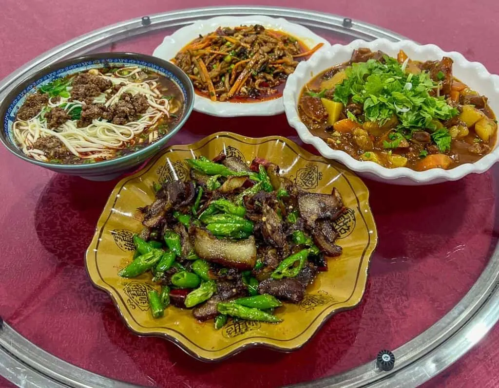 Four dishes of Sichuan food on a red coloured lazy Susan. Dishes are noodles with ground beef, pork and green beans, vegetable stirfry garnished with green herbs and a grated carrot and vegetable stir fry