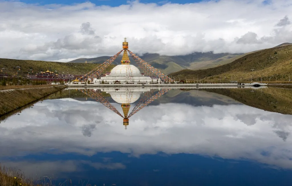 Stupa in Sichuan reflected in a lake with hillsides in the background with a cloudy sky also reflected in the lake