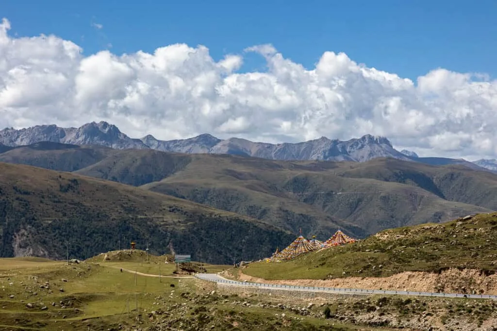 Mountains and green hills with two prayer flag teepees and a curved road in the foreground. The sky is blue with fluffy clouds