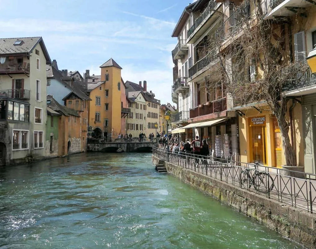 Annecy, France. Looking up one of the canals which is lined with medieval houses painted yellow, peach with railings along a walkway. 
