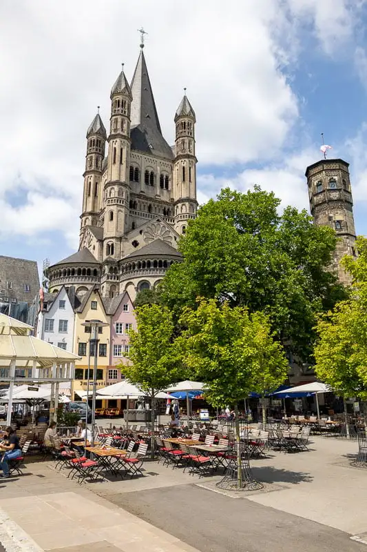 Cologne old town with pastel coloured tall houses and trees in front of an historic towered church. Tables, chairs and parasols sit in front