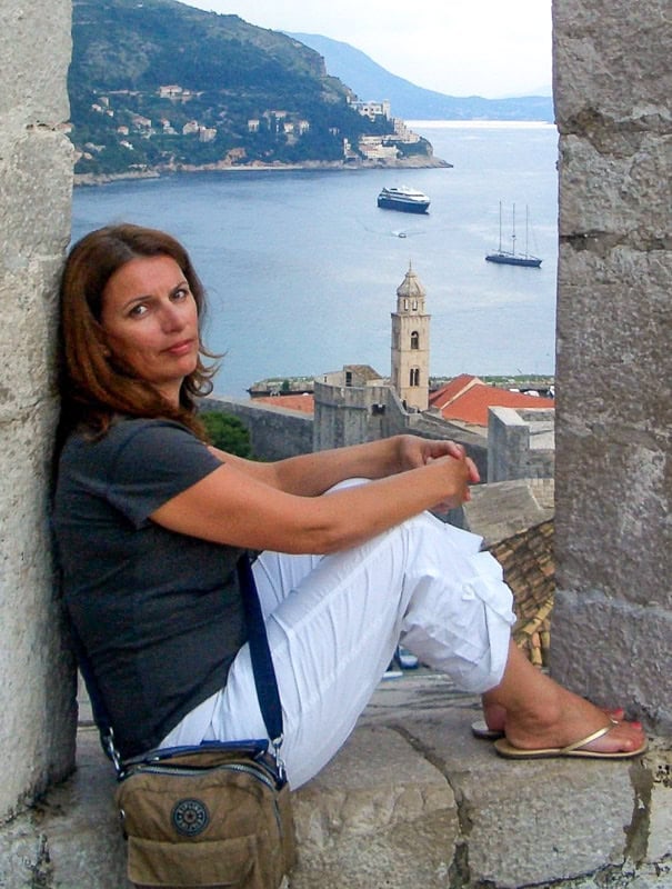 A woman with long brown hair looks at the camera from the stone window she sits on. Behind her is the sea with boats on and a tower and city wall