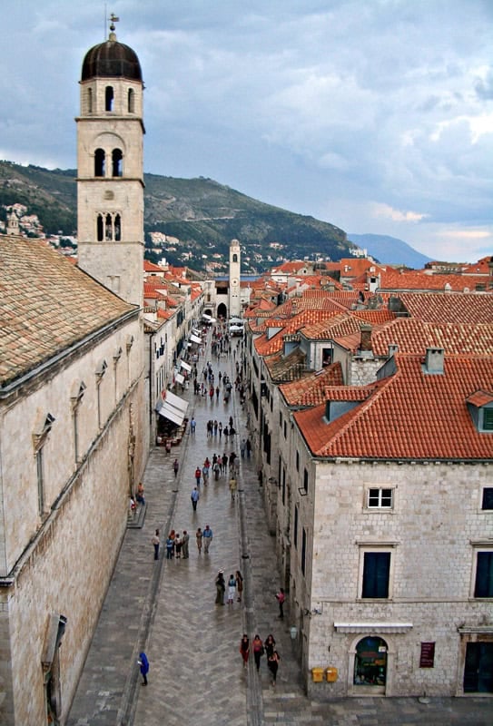 The main marble street in Dubrovnik with the reflections of people on the marble. Red roofed buildings sit on the right and a tower on the left with a hillin the background