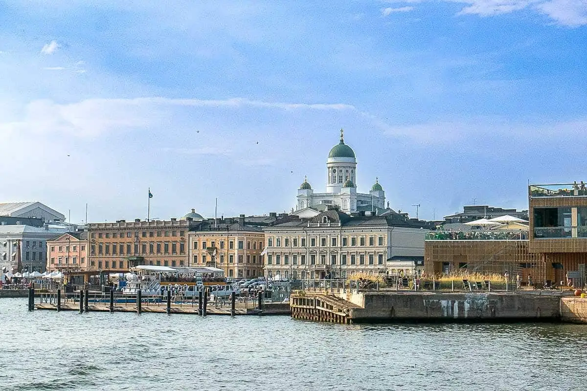 A picturesque view of Helsinki's waterfront featuring historic buildings, a calm sea, and a clear blue sky and the dome of Helsinki Cathedral