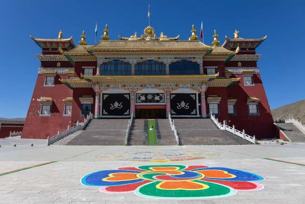 Jiegu Monastery in Yushu, China, features a grand red building with golden roofs, a wide staircase, and a colorful mandala