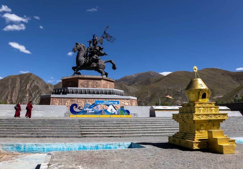 King Gesar Statue in Yushu, China, with a golden chorten, monks, and mountains under a clear sky.