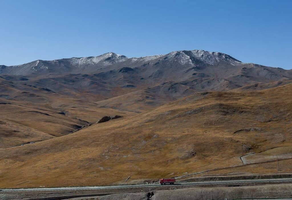 Red truck on a highway through vast, dry hills of Qinghai Province, China, with distant snow-capped mountains.