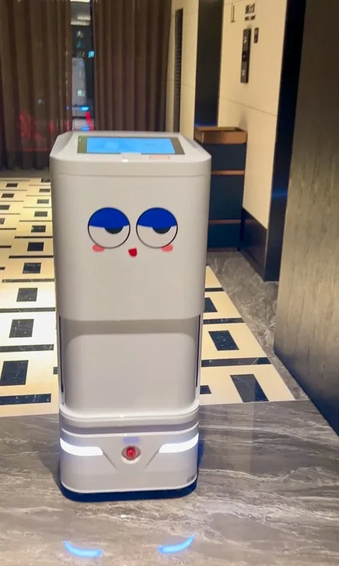 A white room service robot with a cartoon face stands in a modern hotel hallway in China.