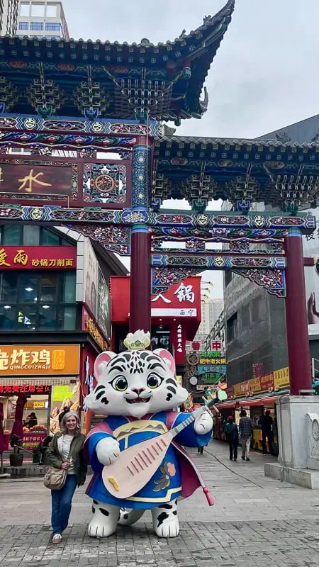 A woman stands beside a snow leopard mascot statue holding a pipa under a traditional archway in Xining, Qinghai.