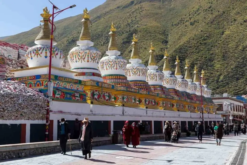 Ornate white and gold stupas line a street with people walking at Jiana Mani Stone City, backed by mountains.